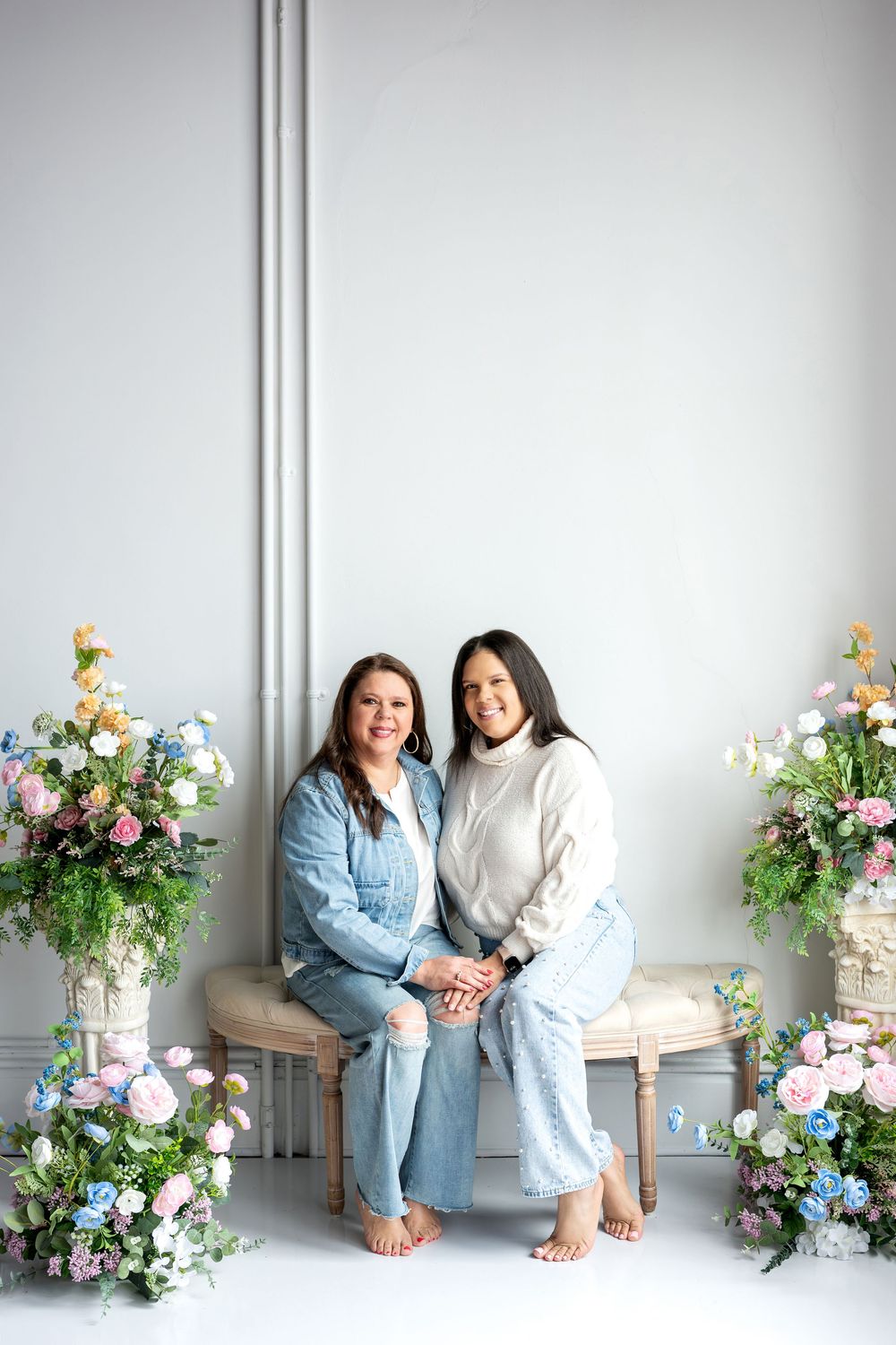 Two barefoot women in jeans sit closely together on bench with colorful floral arrangements.