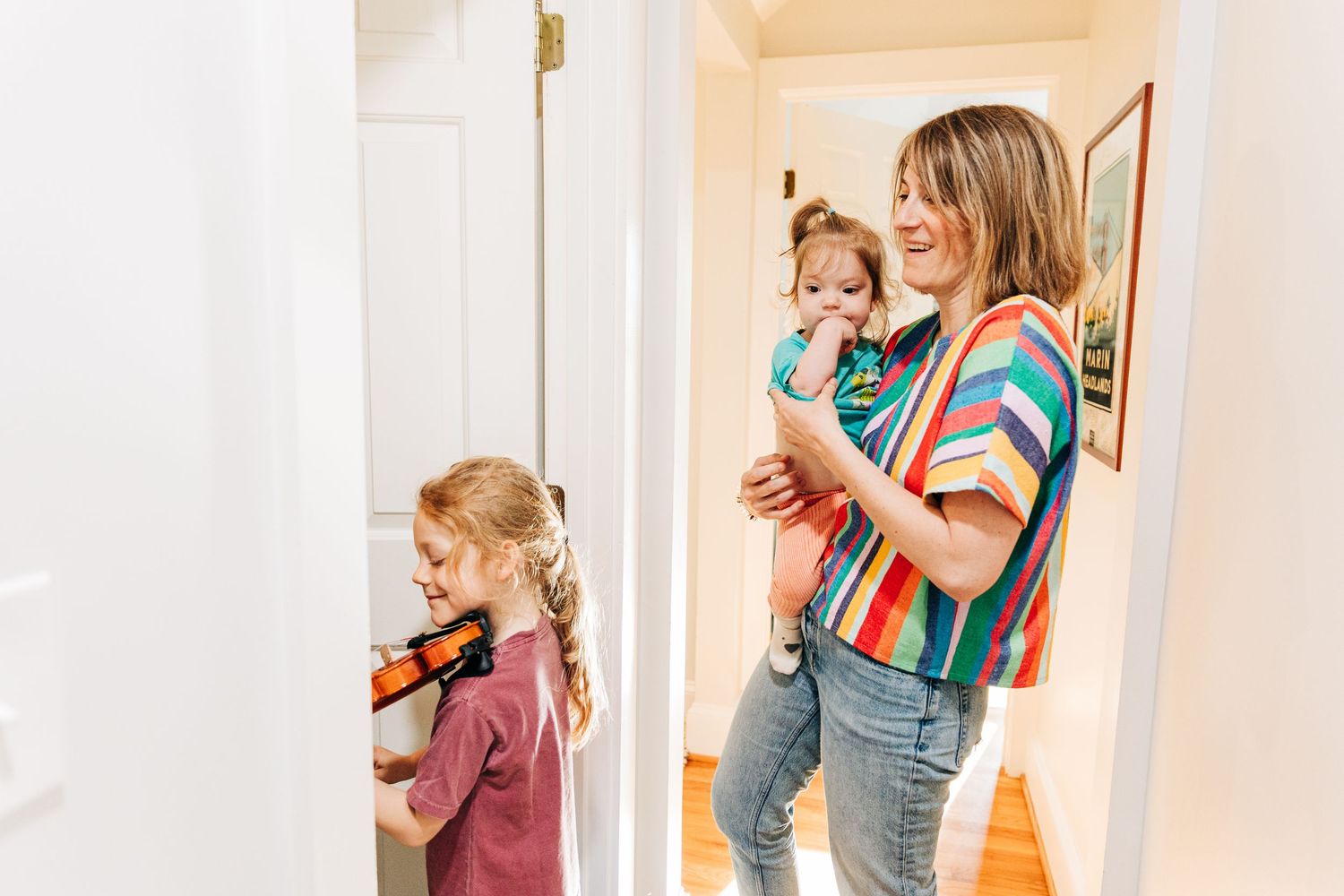A woman holding a toddler watches a young boy play with a violin in a bright home hallway in Charlotte, NC