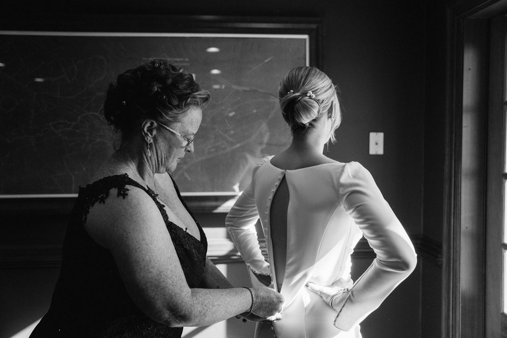 Black and white photo of a woman helping a bride button her elegant wedding dress before the ceremony.