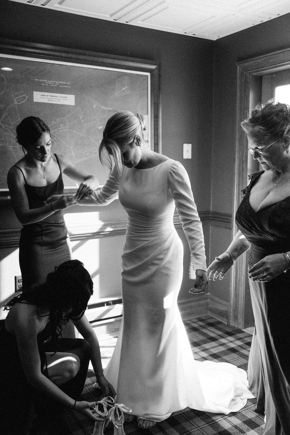 Black and white photo of bride in elegant gown being helped by bridesmaids and mother before wedding ceremony.