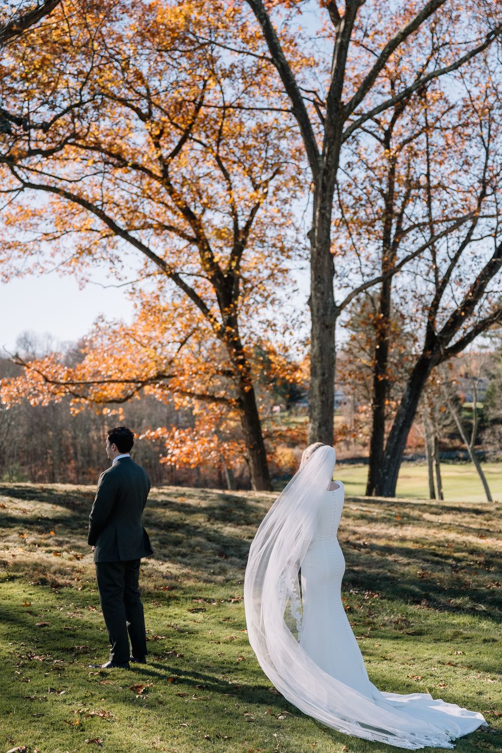 Bride in cathedral veil and groom stand apart for first look on autumn lawn.