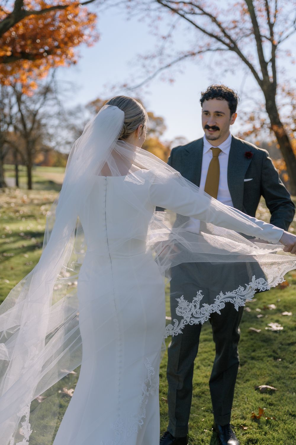 Bride twirling her lace-trimmed veil while groom smiles during outdoor fall wedding photos.