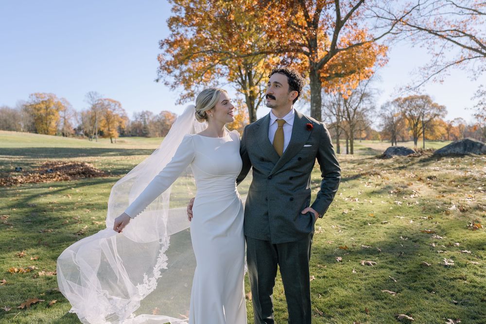 Bride and groom share a joyful moment outdoors in autumn, her lace veil flowing in the breeze.