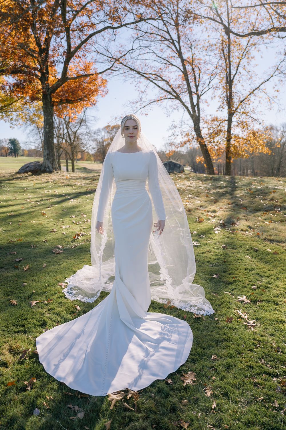 Elegant bride in a sleek white long-sleeve gown and lace cathedral veil stands in an autumn park.