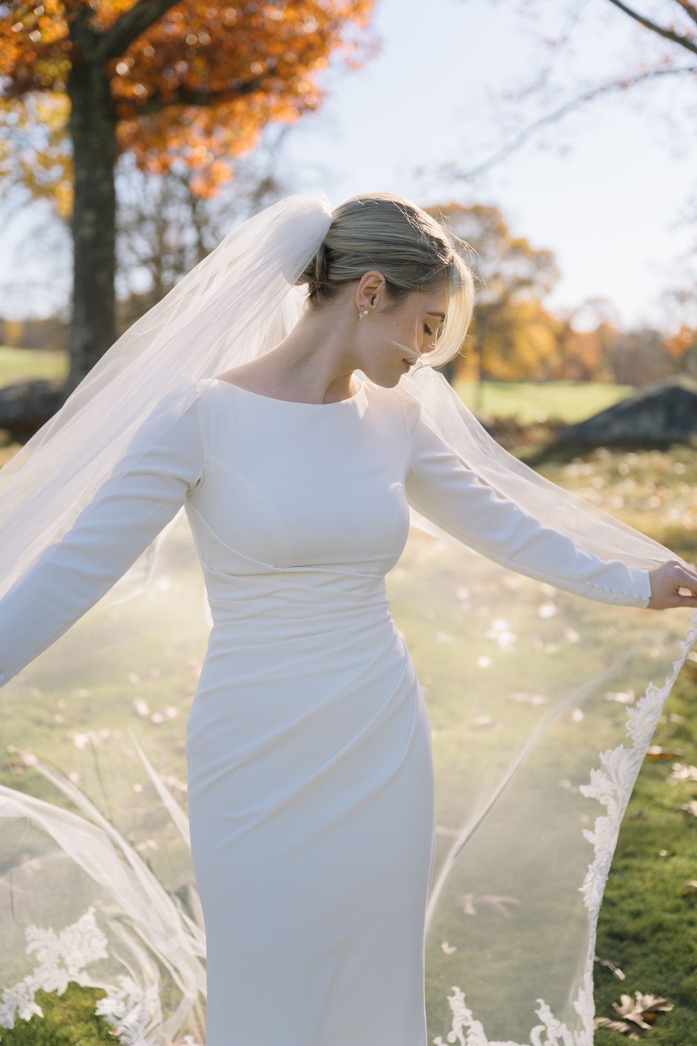 Bride in a fitted white long-sleeve wedding dress holds her flowing lace veil in golden autumn light.