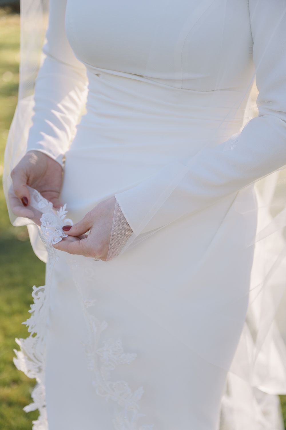 Close-up of a bride in a long-sleeve white wedding gown holding delicate lace details outdoors.