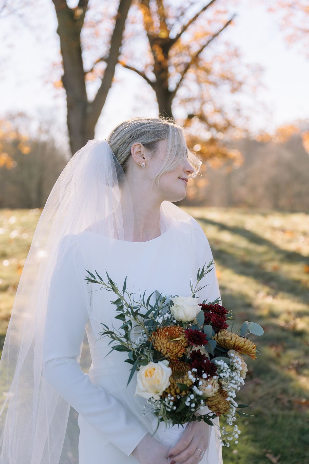 Bride in white long-sleeve gown holding autumn bouquet with red, yellow flowers, wearing veil outdoors.