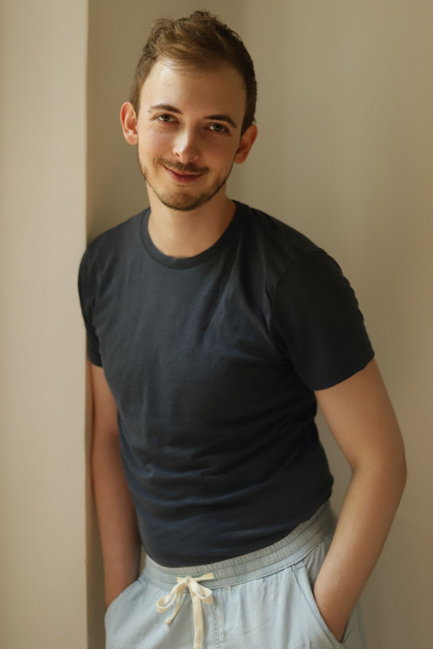 Young man smiling in black t-shirt and light gray drawstring shorts, leaning against a wall indoors.