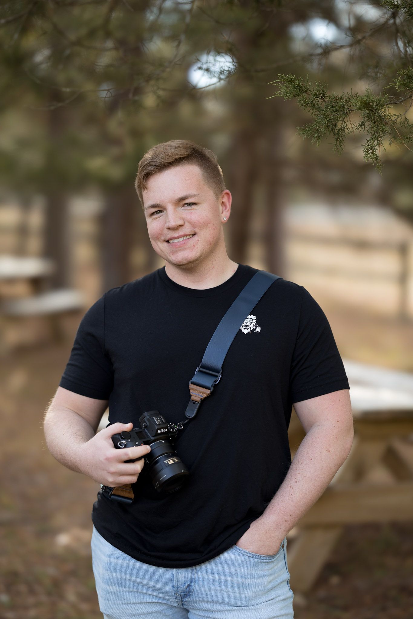 Young man in black t-shirt holding a DSLR camera with strap, smiling outdoors in a pine tree forest setting.