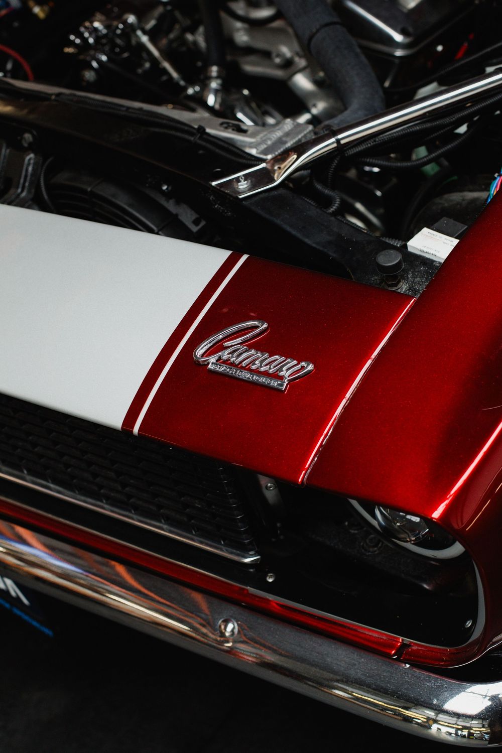 Close-up of a classic red Chevrolet Camaro with white racing stripes and chrome badge, hood open revealing engine.