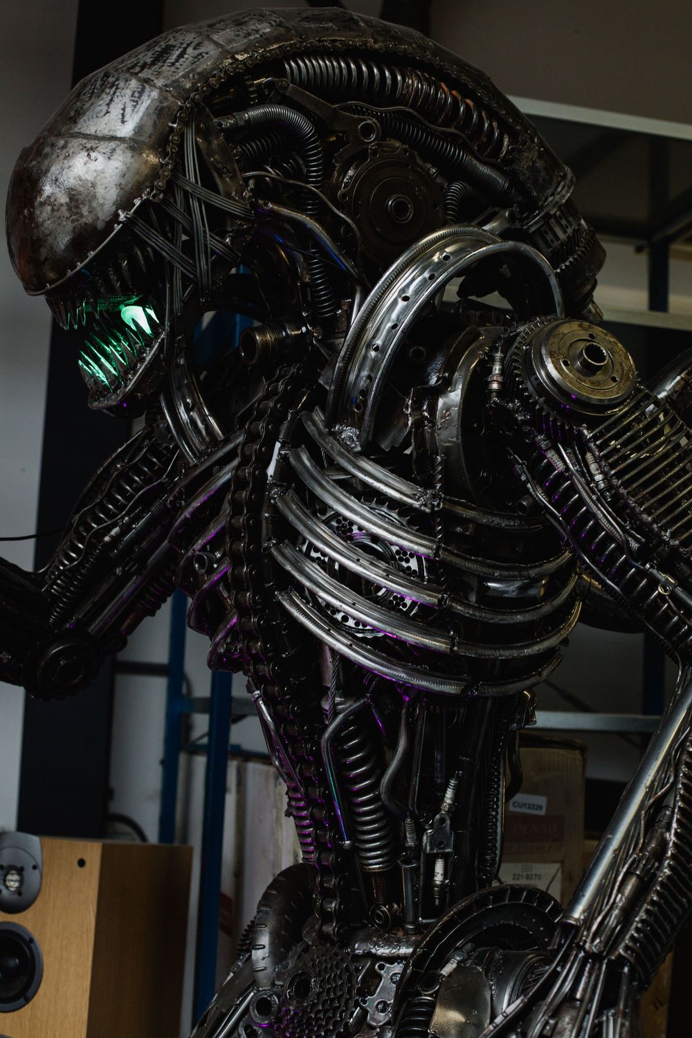 Intricate life-size alien sculpture crafted from recycled automotive parts, gears, and metal components with glowing green eyes.