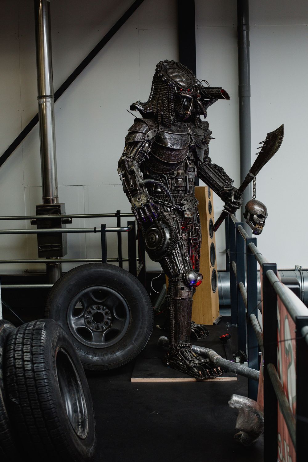Full-body Predator sculpture made from scrap metal and car parts, holding a spear, displayed in a garage setting.