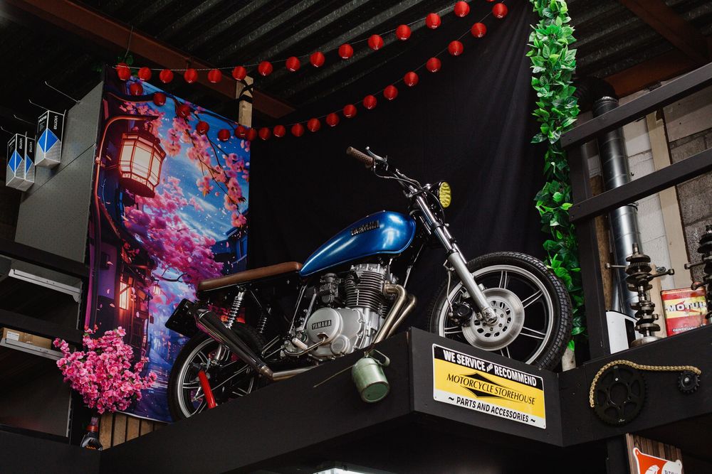 Blue vintage motorcycle mounted on a shelf above tires, decorated with cherry blossom artwork and red lanterns indoors.