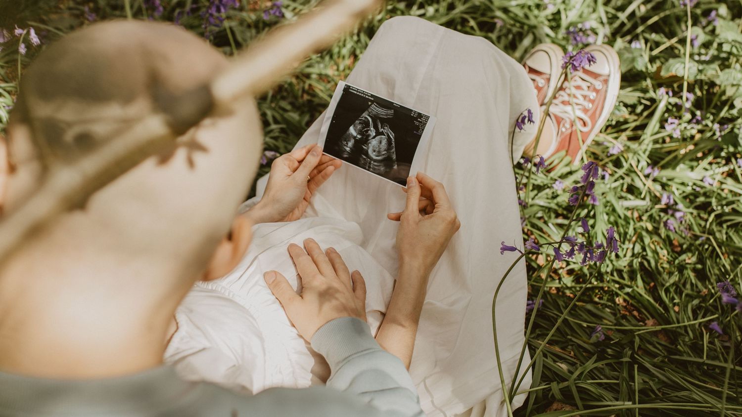 Couple sitting in grass holding ultrasound photo over pregnant belly, surrounded by wildflowers.
