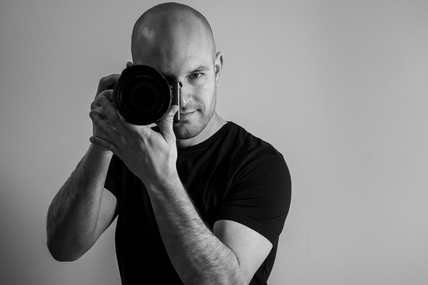 Professional photographer wearing a black t-shirt holds a camera up to their face in a black and white portrait shot.