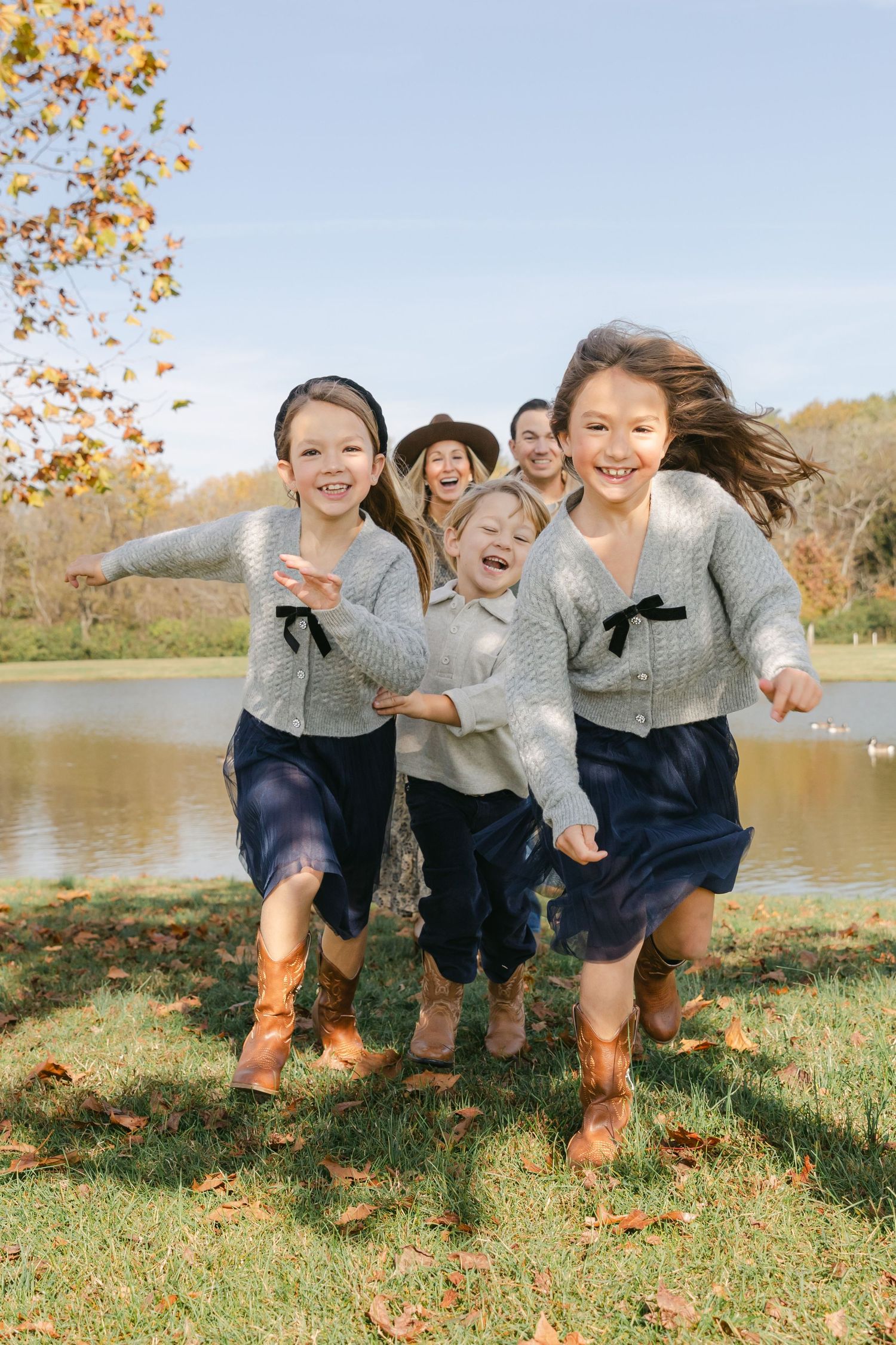 A joyful family with children runs along a farm path with white fences and a green-roofed barn in the background.