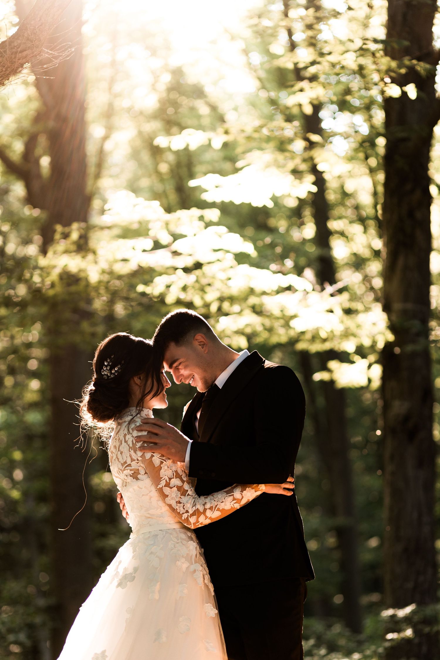 A bride and groom share a joyful embrace surrounded by sunlit trees in a forest setting.