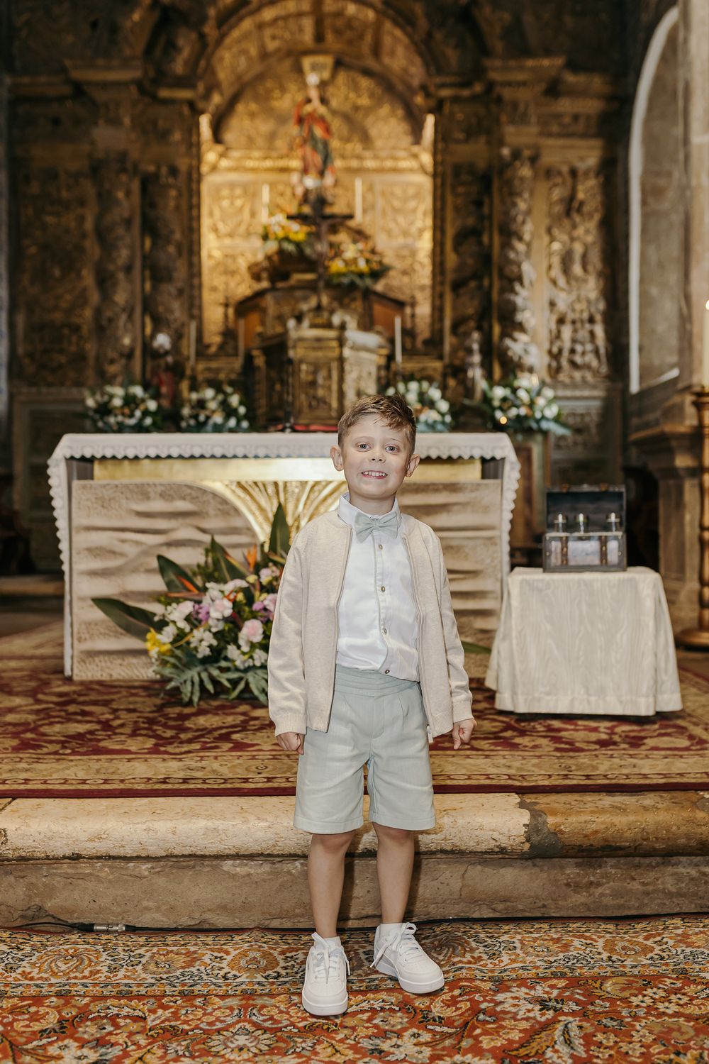 Young boy in beige jacket and mint shorts stands before ornate golden church altar with floral arrangements. Jo Matos Fotografia Reportagem