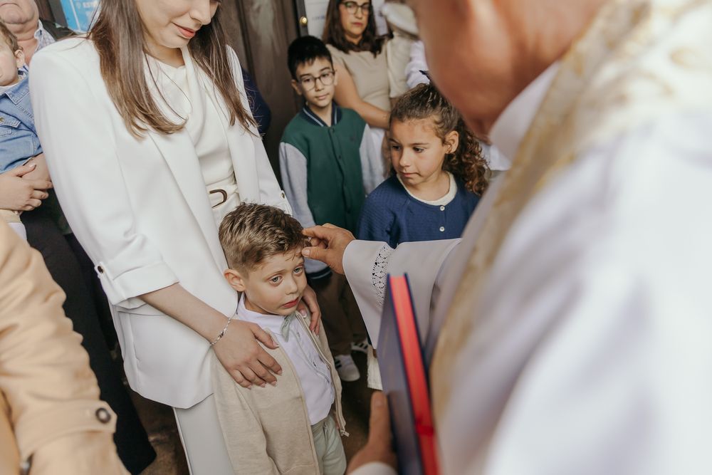 A young boy in white stands with a woman during a religious ceremony with children and a priest.