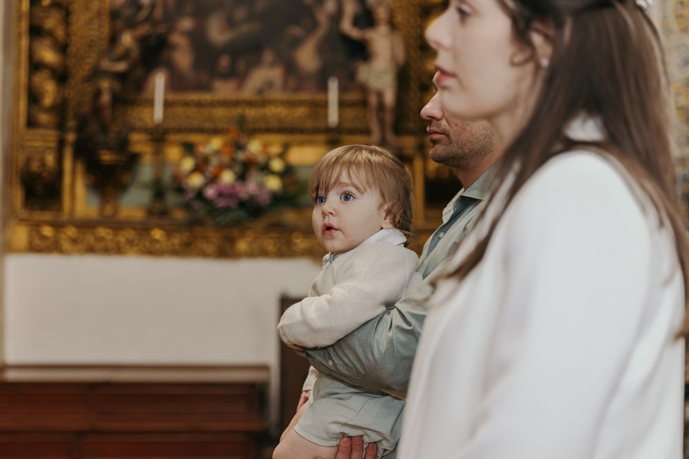 A woman holds a baby in a church with a golden ornate altar in the background.Jo Matos Fotografia Reportagem de Batismo