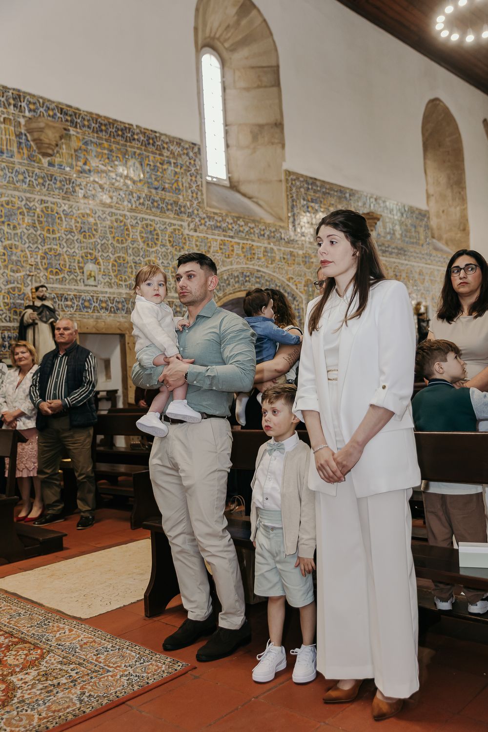Family standing in a decorated church during a baptism ceremony, with ornate blue tile walls in the background.Jo Matos Fotografia Reportage