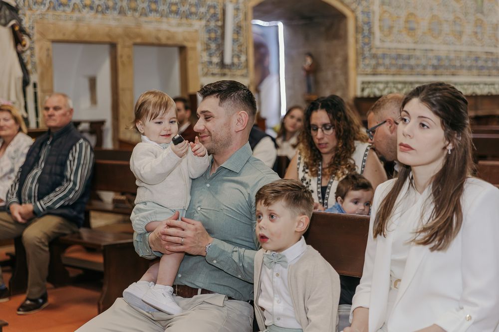 Family sitting in church pews during a baptism ceremony, with a man holding a toddler and a woman beside two children.Jo Matos Fotografia Re