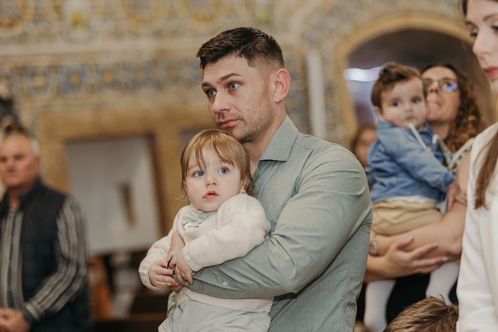 A man in a light blue shirt holds a baby in white at a church baptism ceremony.Jo Matos Fotografia Reportagem de Batismo