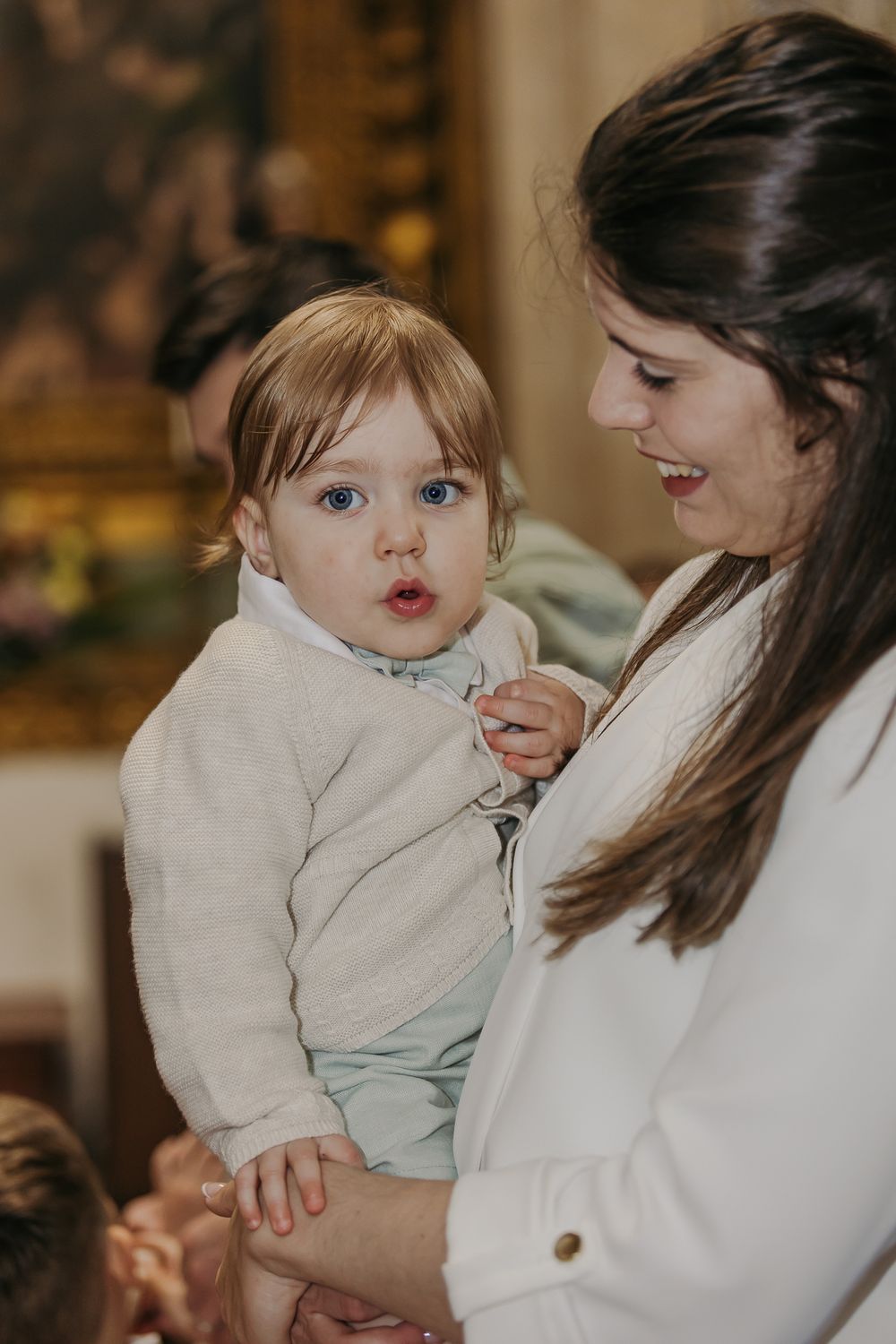 A young woman in white holds a wide-eyed baby in a beige outfit at an elegant indoor event.Jo Matos Fotografia Reportagem de Batismo