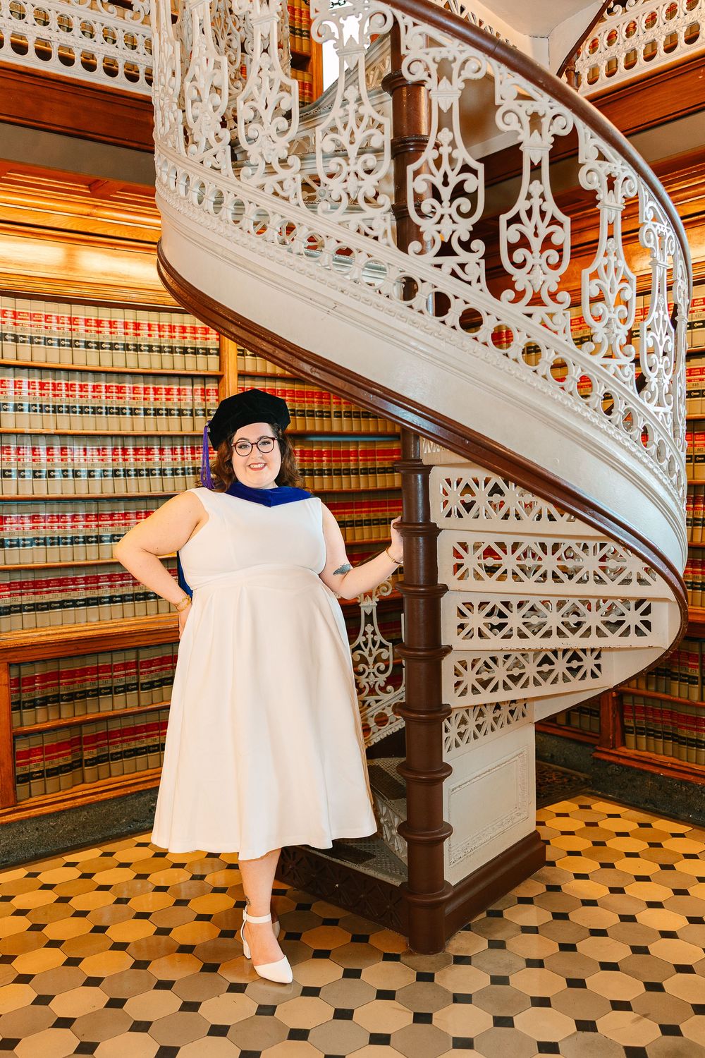 A woman in a white dress and graduation cap smiles with her hand on her hip in front of shelves of library books and a spiral staircase.