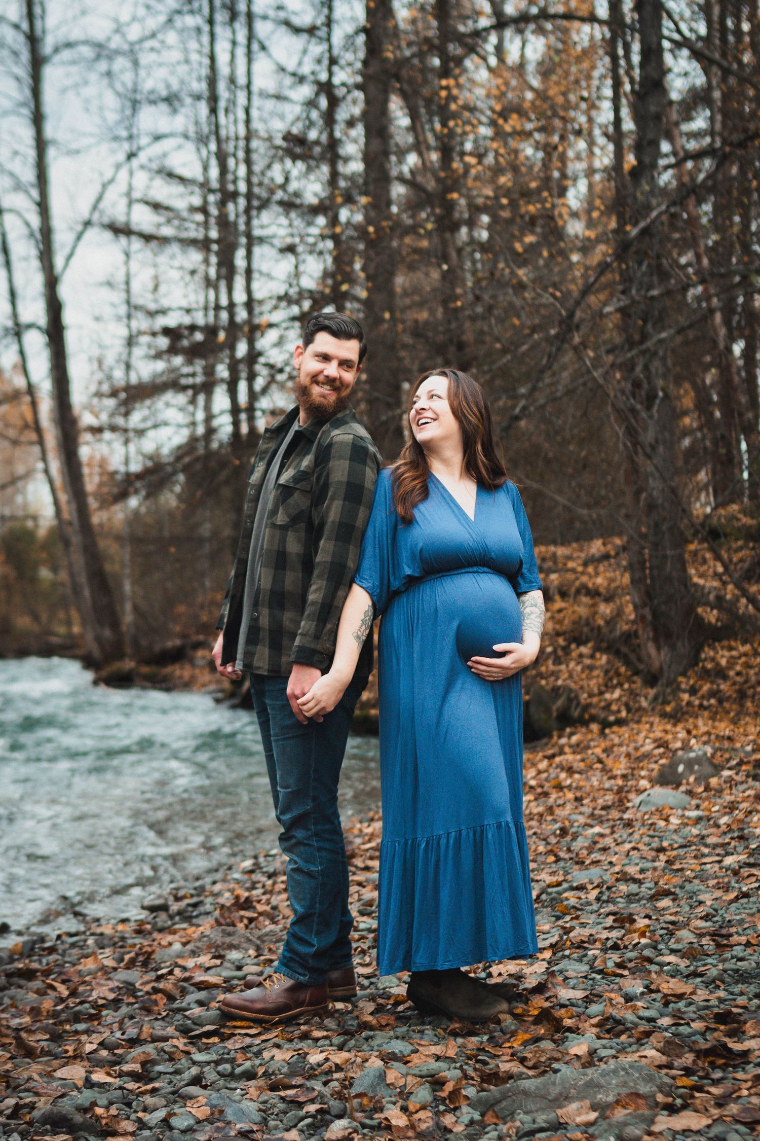 Couple holding hands by a river in fall, pregnant woman in blue dress cradling belly, man in plaid shirt smiling outdoors.