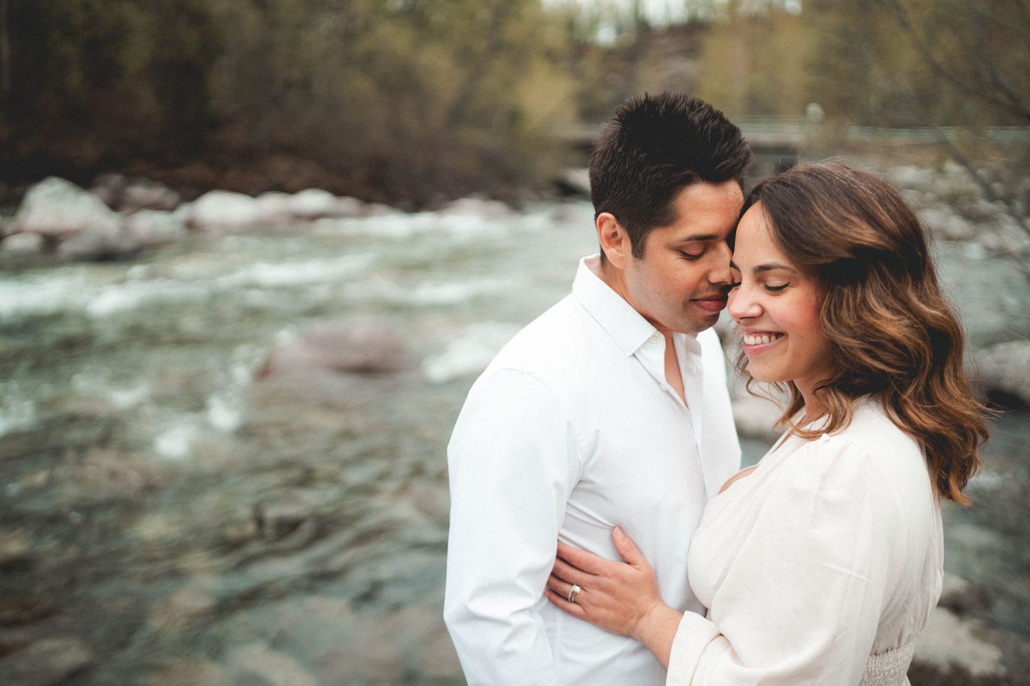 Couple embracing romantically by a rocky river, wearing white outfits, smiling tenderly in a natural outdoor setting.