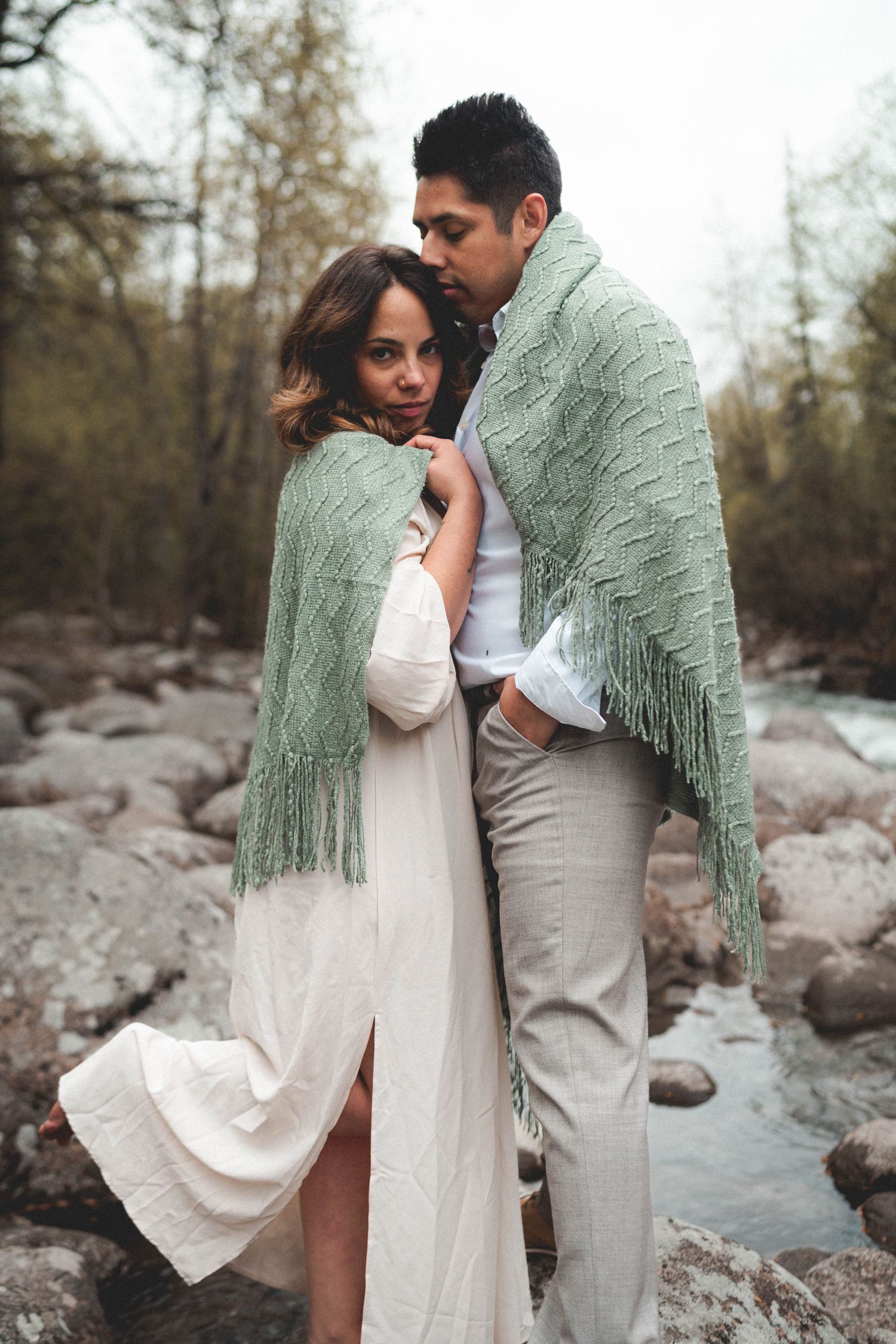Couple wrapped in green knit shawl embracing on rocky riverbank surrounded by forest trees.