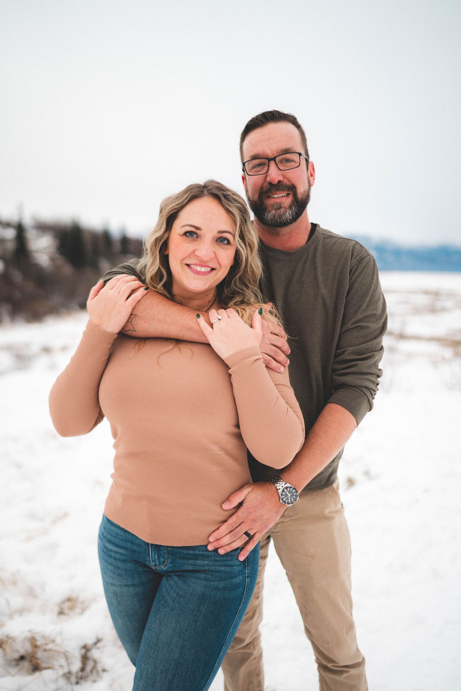 Couple embracing in snowy winter landscape, smiling at camera, woman in pink sweater and jeans, man in olive shirt with glasses.