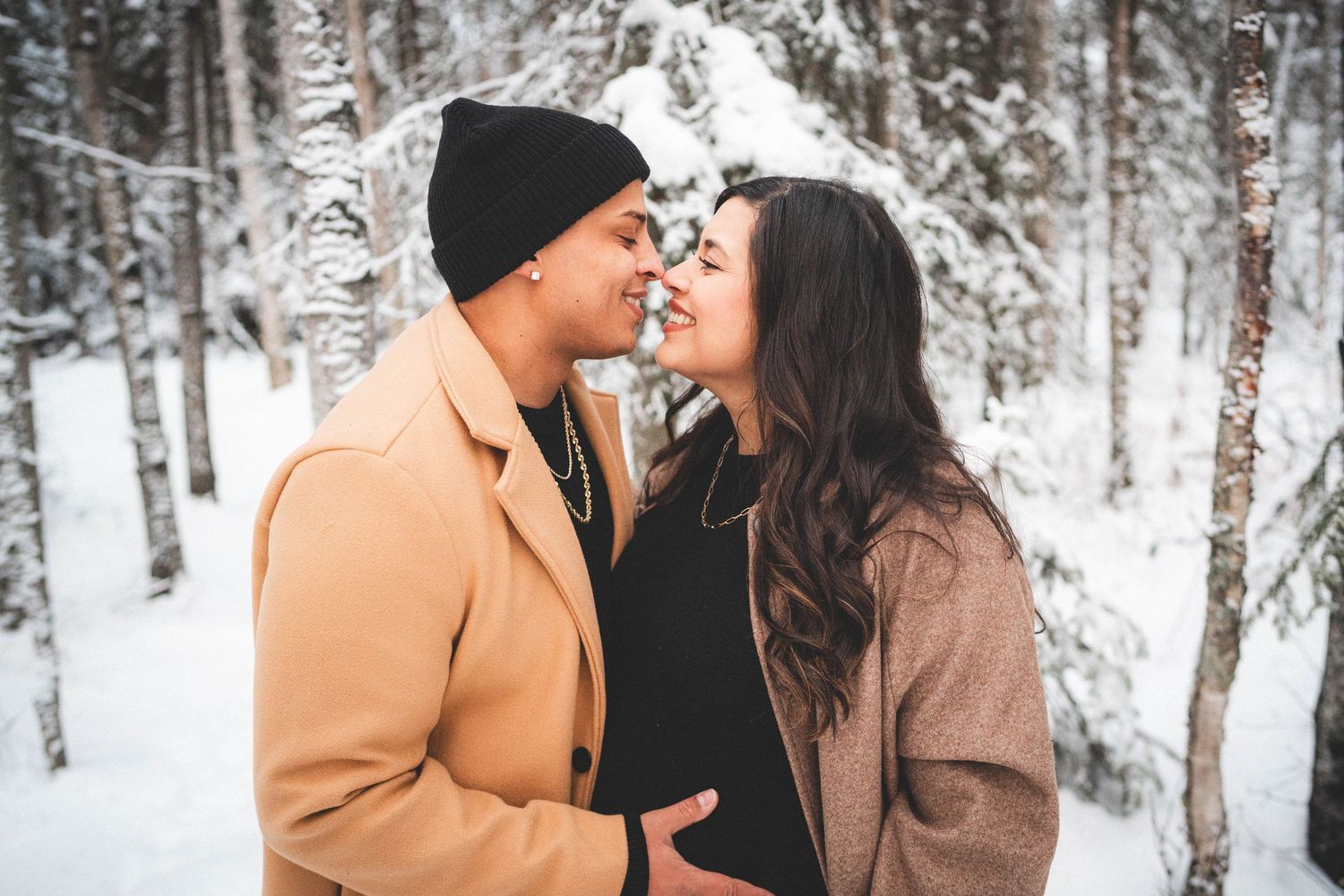Couple sharing a romantic moment in a snowy winter forest, woman showing a baby bump in matching beige coats.
