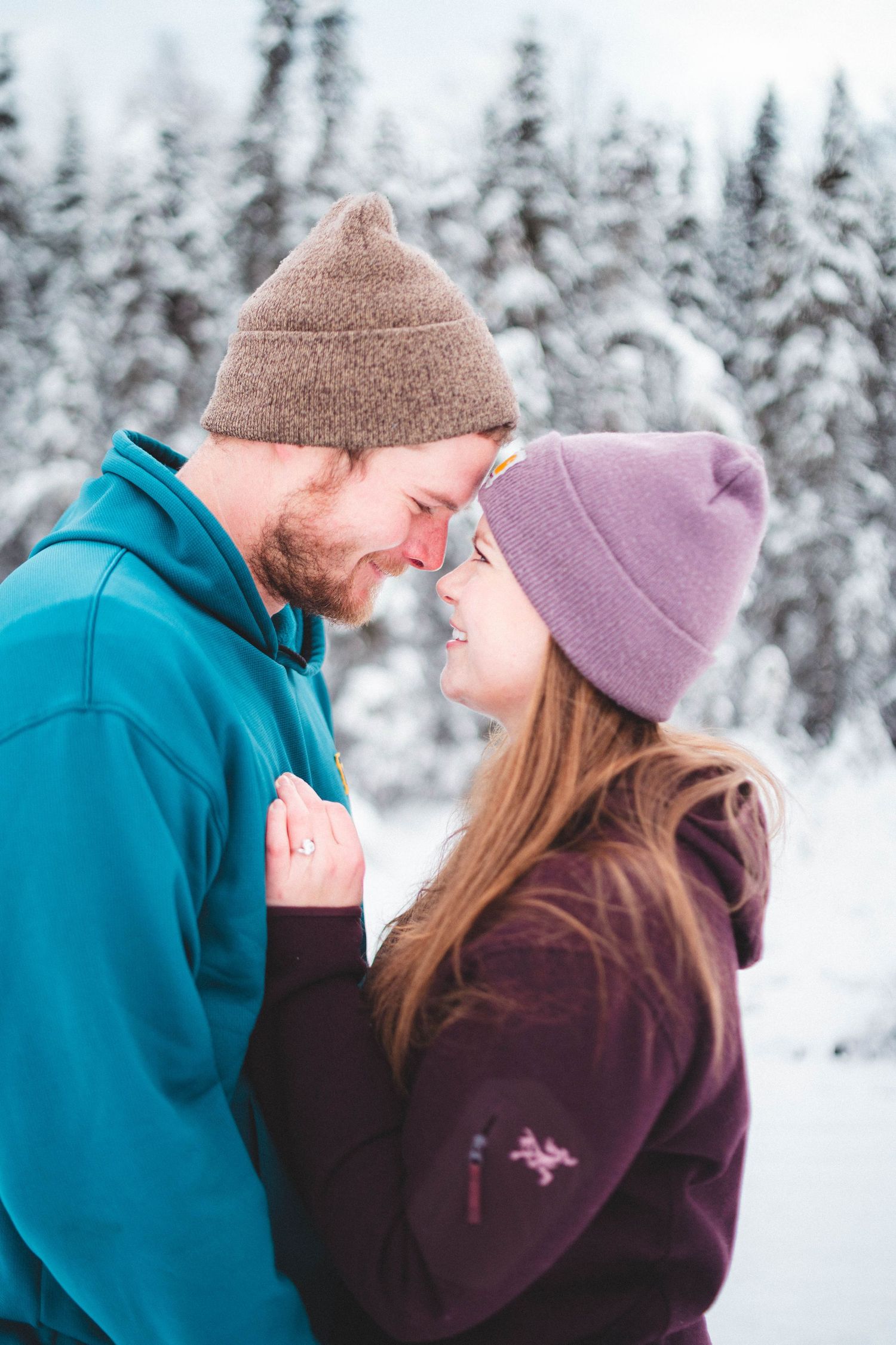 Couple in winter hats smiling forehead-to-forehead in snowy forest, wearing teal and purple fleece jackets.