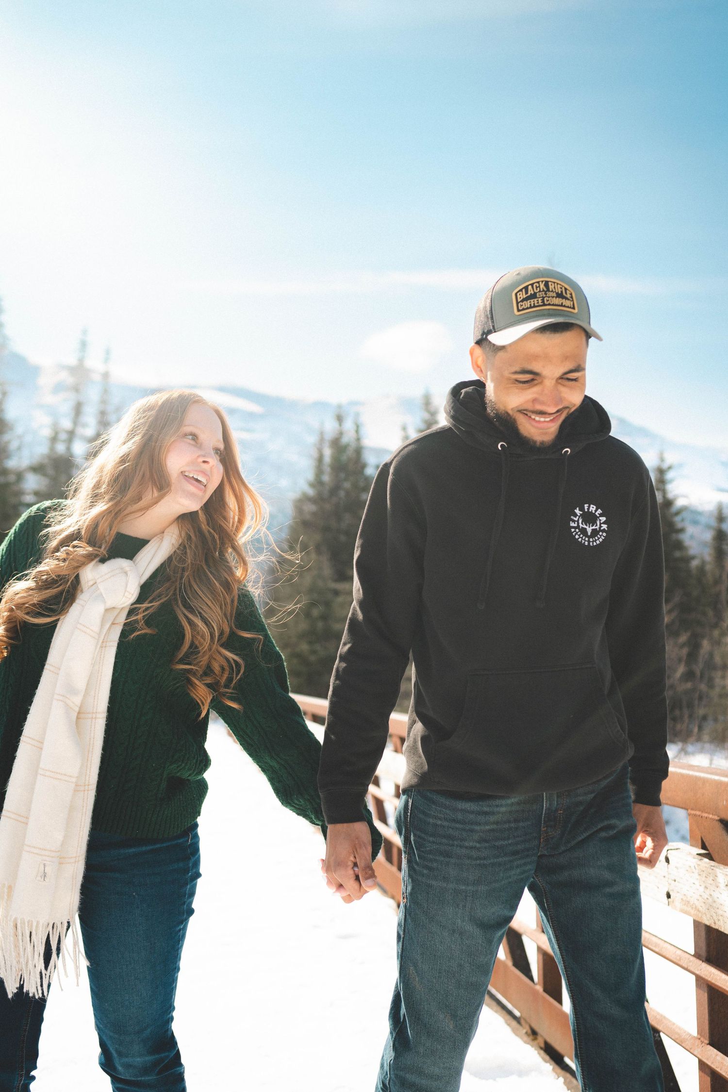 Couple holding hands and laughing in snowy mountain landscape, wearing winter clothes on a sunny day.