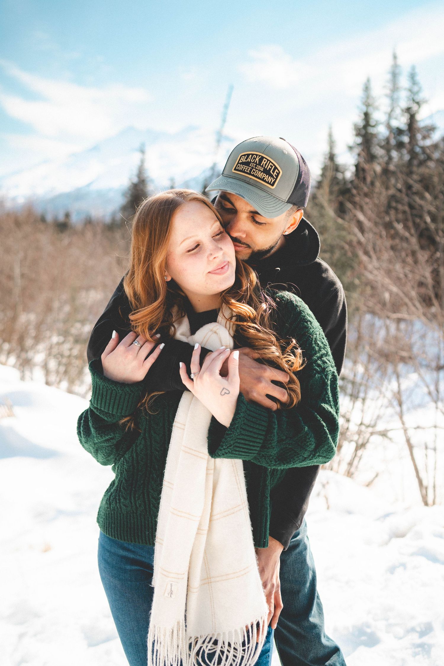 Couple embracing in snowy winter landscape, woman in green sweater and white scarf, man in cap kissing her cheek.