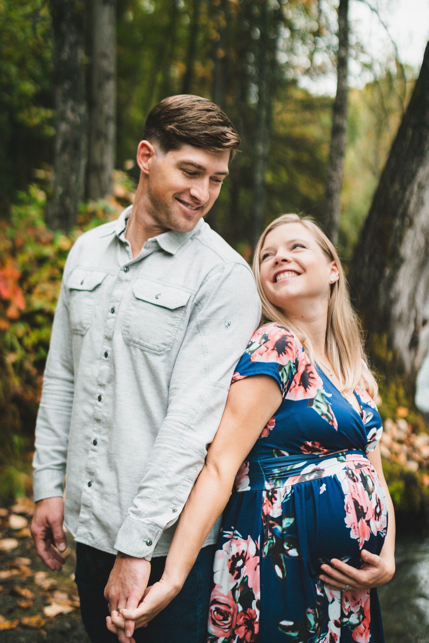 Smiling pregnant couple holding hands outdoors in autumn forest, woman in floral blue dress cradling baby bump.