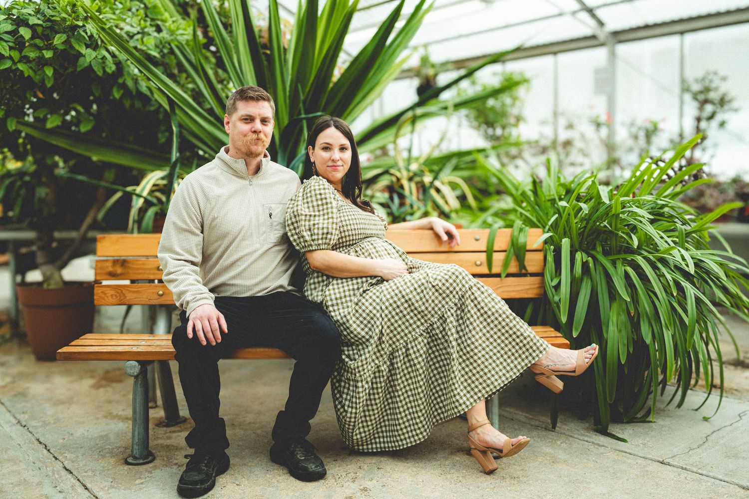 Couple posing on a wooden bench surrounded by lush tropical plants in a greenhouse setting.