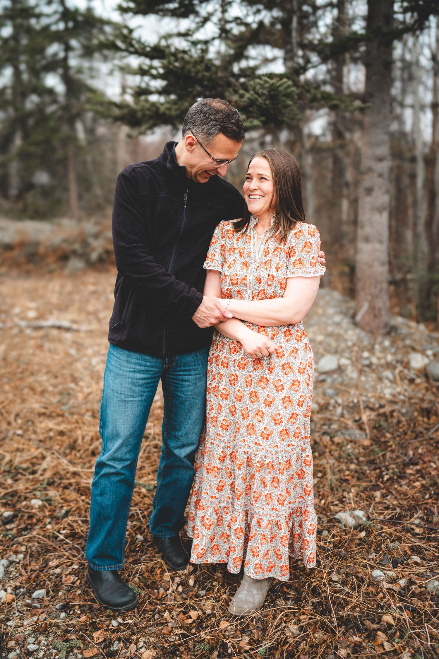Couple holding hands and smiling in a wooded outdoor setting, woman in floral dress, man in black jacket and jeans.