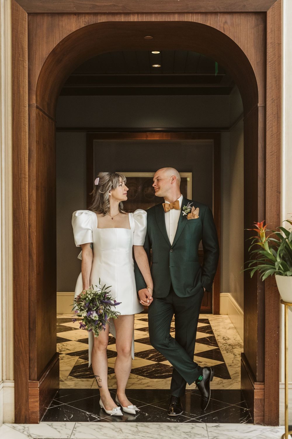Bride in white mini dress and groom in dark green suit hold hands under wooden arch at elegant wedding venue.