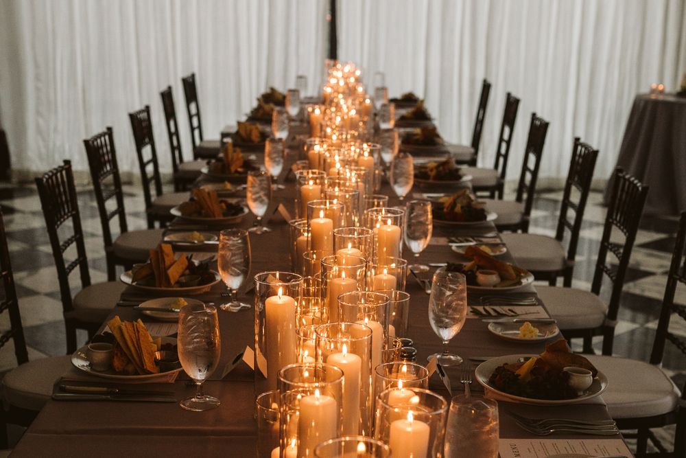 Elegant candlelit wedding reception table with glowing pillar candles, wine glasses, and dark wood chairs in a draped venue.