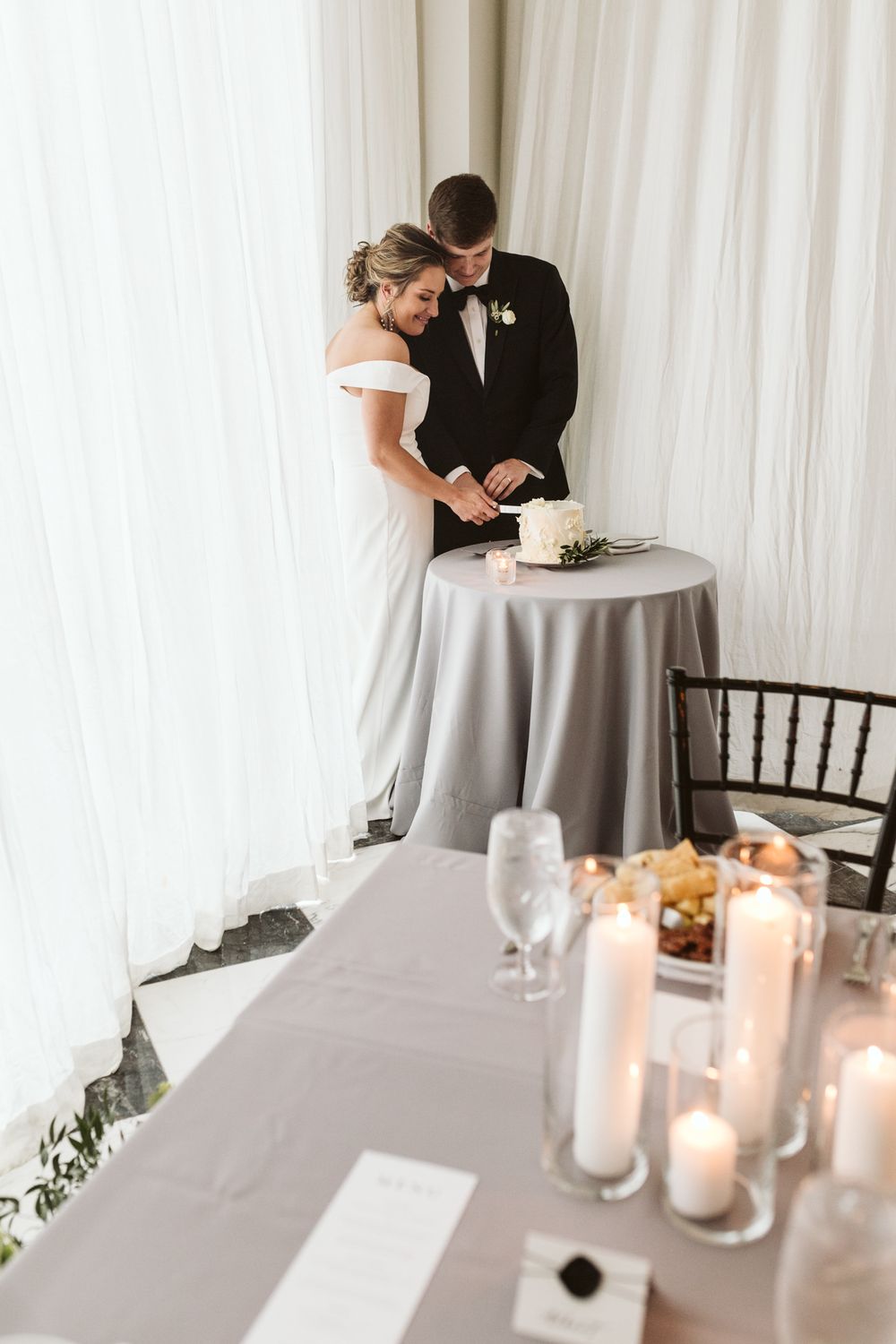 Bride and groom cutting their wedding cake at a reception with candles and gray linen decor.