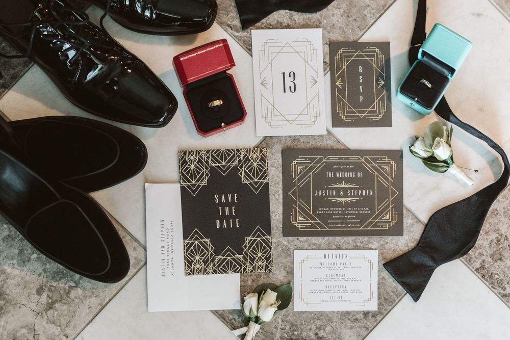 Art deco wedding stationery suite with save the date cards, invitations, rings, bow tie, and black dress shoes on marble surface.