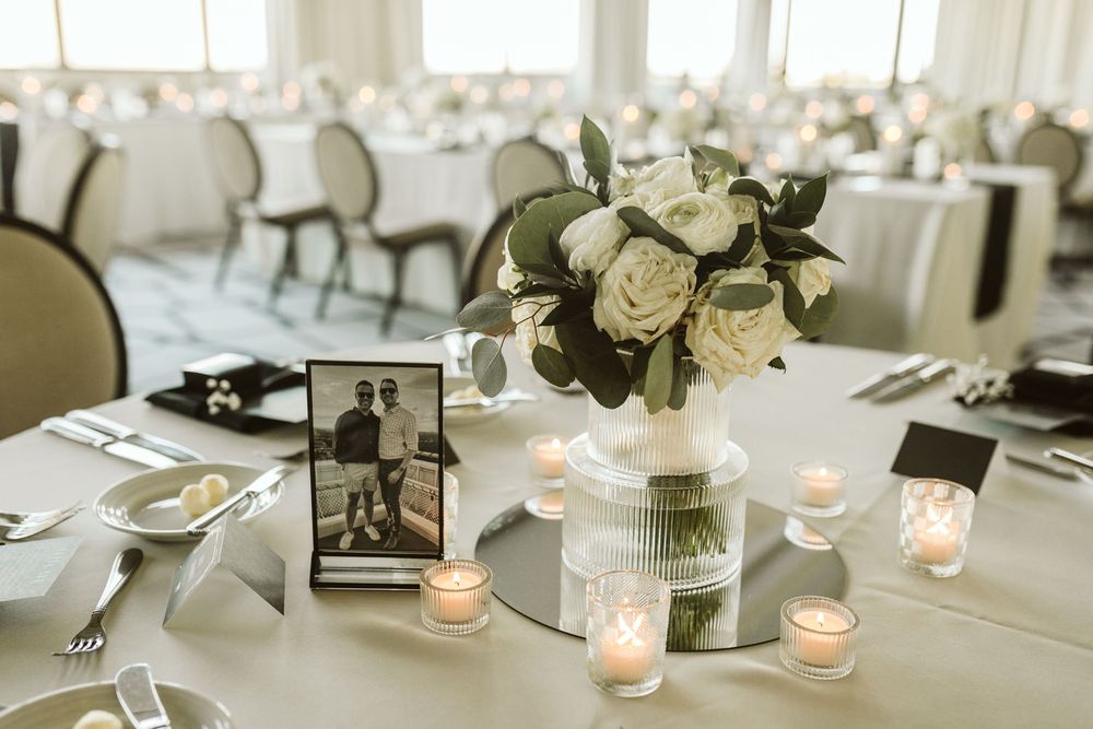 Elegant wedding reception table with white roses, greenery centerpiece, candles, and a framed couples photo on white linen.