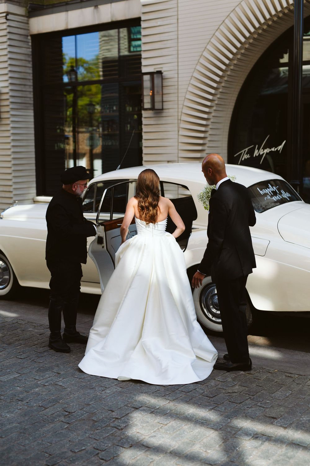 Bride in white ball gown entering vintage cream Rolls-Royce wedding car, assisted by two attendants outside elegant venue.