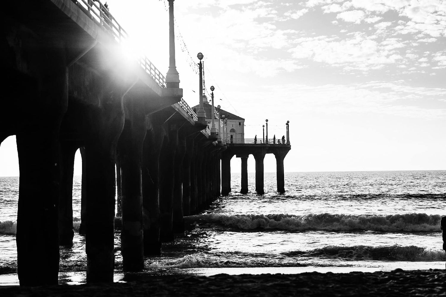 Silhouetted pier extends into ocean waves with dramatic sunburst filtering through pillars in black and white.