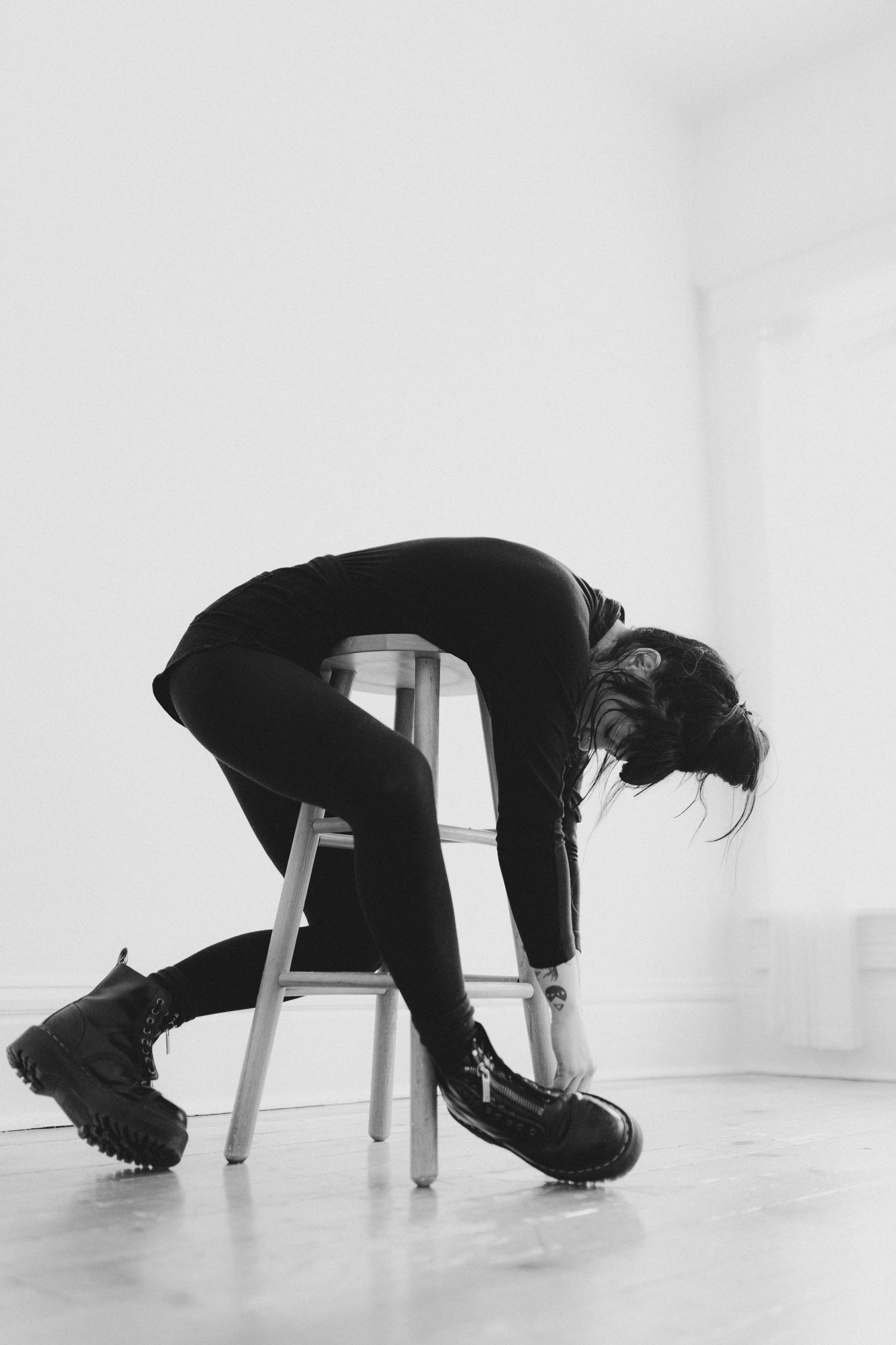 Figure in black clothing bends over metal stool in minimalist white room.