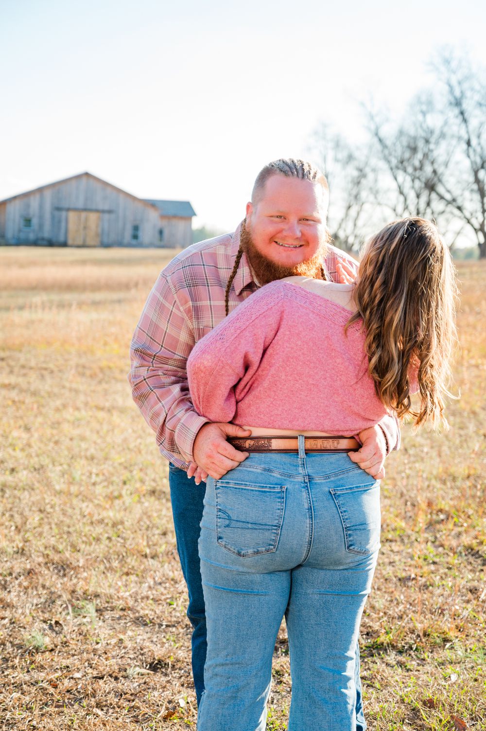 Couple embracing in a golden field near a rustic barn on a sunny autumn day, man smiling warmly.