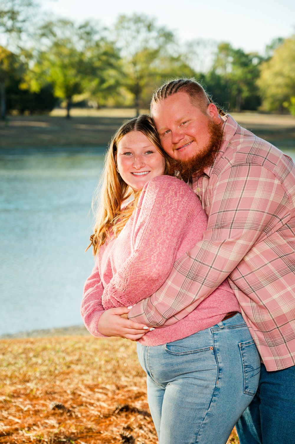 A smiling couple in matching pink outfits embracing outdoors near a lake with autumn foliage in the background.
